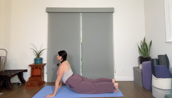 A woman in loose purple pants and a gray tank practices Upward Facing Dog on a blue mat