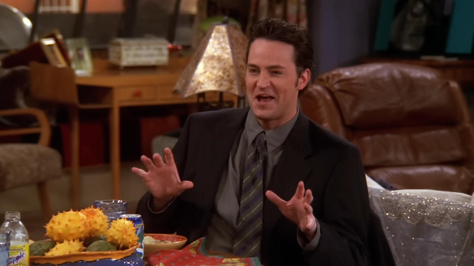 Matthew Perry smiles and gestures with his hands while sitting indoors in a living room setting