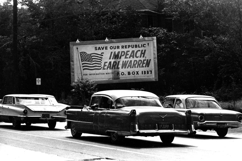 A highway with old cars on it and a billboard that says 'IMPEACH EARL WARREN' on the side.