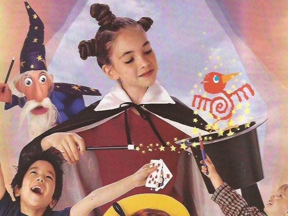 Photo of India Kushner with her brown hair pulled up into three buns, one on the top of her head and two on either side. She wears a magician's cape and points a wand at an upside-down top hat, which she holds in her other hand. She smiles, looking down at the hat, out of which pops a cartoon bird with stars around it. Below her is a blonde girl wearing a plaid dress and pointing a wand up at India. To the left of that is a boy with black hair and dark eye raising his arms and cheering, holding playing cards in his left hand. Over India's right shoulder is a magician puppet. 