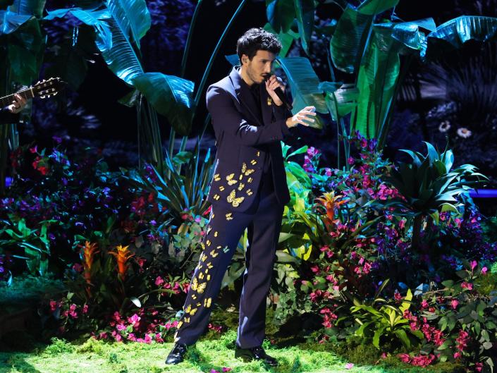 Sebastian in a dark blue-purple suit with yellow butterfly appliques going up his right pant leg, bottom of the right side of his jacket, and top of the left side of this jacket.