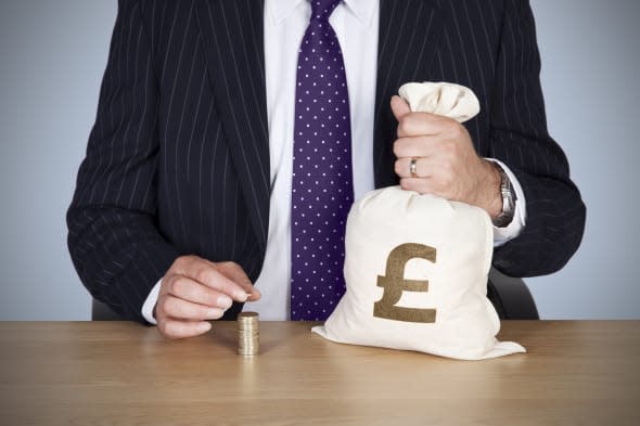 A British banker sat at a desk holding a money bag and adding a pound coin to a stack of coins.