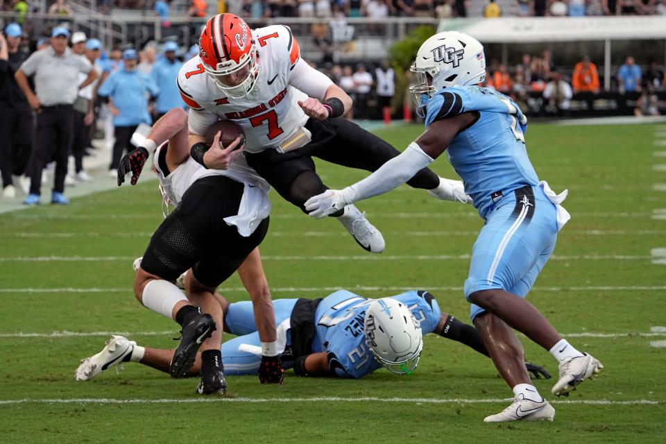 Oklahoma State quarterback Alan Bowman (7) leaps for extra yardage over Central Florida defensive back Nikai Martinez (21) and linebacker Jason Johnson, right, during the first half of an NCAA college football game, Saturday, Nov. 11, 2023, in Orlando, Fla. (AP Photo/John Raoux)