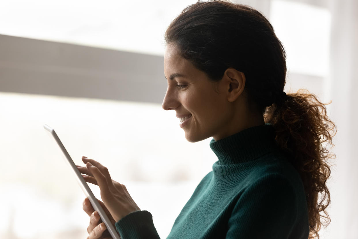 Side view of smiling casual hispanic woman holding electronic tablet computer surfing social networks learning virtual course reading e book. Calm young female using tab pc by window