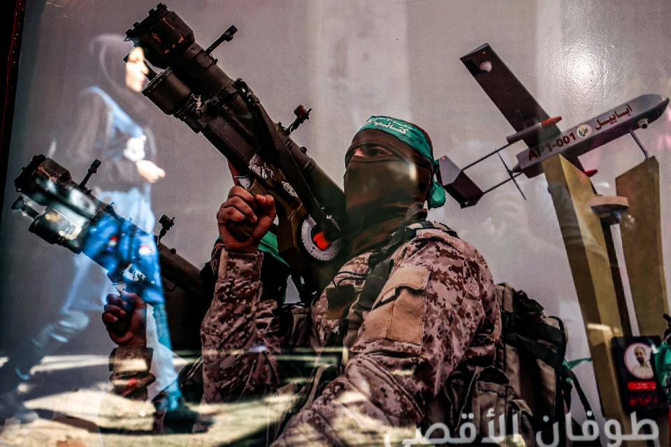 A reflection shows a woman walking past a poster in a shop window showing a Hamas fighter.