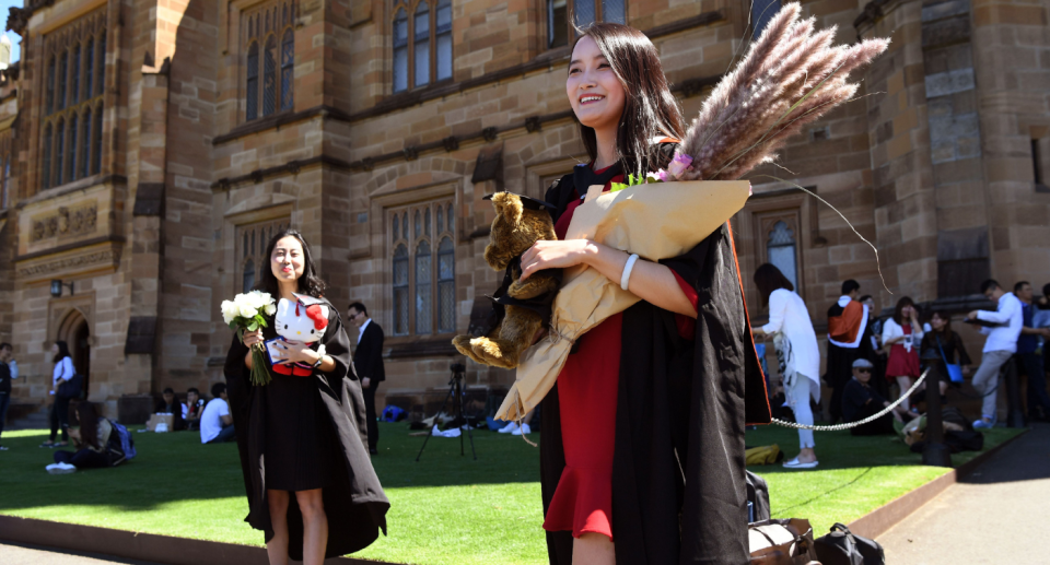 Two graduates stand smiling out the front of a sandstone university.