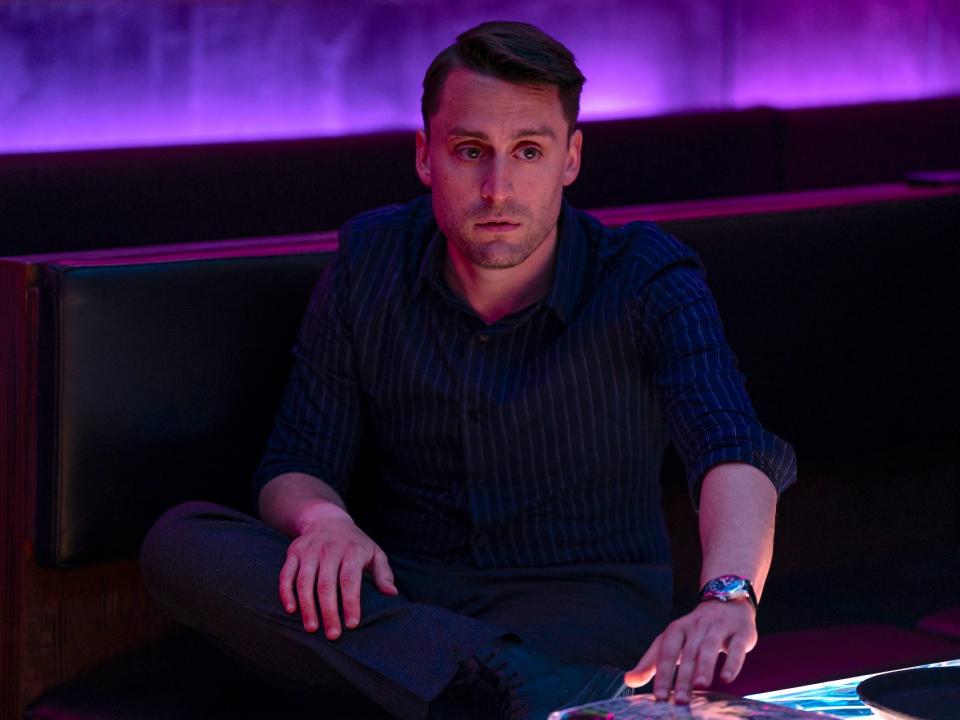 A white man wearing dress clothes and looking upset in a purple-lit lounge room.