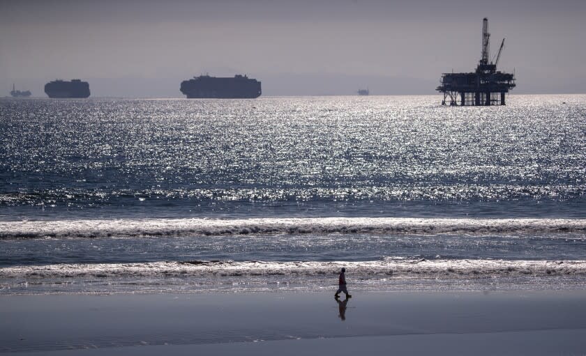 Huntington Beach, CA - October 05: Container ships and an oil derrick line the horizon as a lone environmental oil spill cleanup crew member searches the beach, cleaning up oil chucks from a major oil spill in Huntington Beach Tuesday, Oct. 5, 2021. Environmental cleanup crews are spreading out across Huntington Beach and Newport Beach to cleanup the damage from a major oil spill off the Orange County coast that left crude spoiling beaches, killing fish and birds and threatening local wetlands. The oil slick is believed to have originated from a pipeline leak, pouring 126,000 gallons into the coastal waters and seeping into the Talbert Marsh as lifeguards deployed floating barriers known as booms to try to stop further incursion, said Jennifer Carey, Huntington Beach city spokesperson. At sunrise Sunday, oil was on the sand in some parts of Huntington Beach with slicks visible in the ocean as well. "We classify this as a major spill, and it is a high priority to us to mitigate any environmental concerns," Carey said. "It's all hands on deck." (Allen J. Schaben / Los Angeles Times)