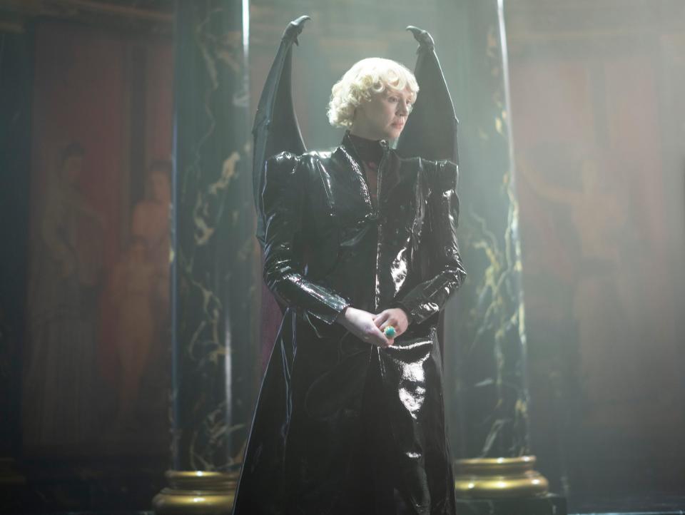 gwendoline christie as lucifer morningstar in the sandman. she's a tall woman with short, curled blonde hair. she's wearing an a-line leather dress with prominent shoulders, and has two leathery black wings on her back. there's a large turquoise ring on her left hand.