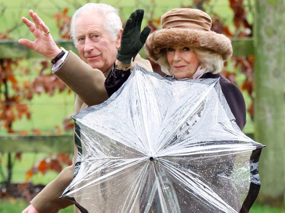 King Charles and Queen Camilla wave while holding an umbrella.
