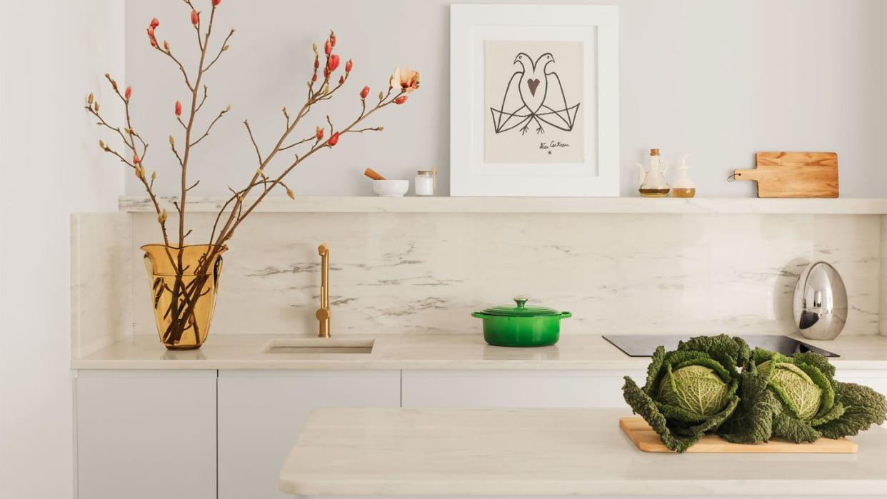 in the kitchen, an island and counter, with a built in sink, are topped with portuguese rosa marble, on the counter is a vase with tall salmon colored buds and a green saucepot, on a shelf is a drawing of two birds