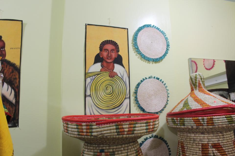 The art in the background depicts a woman weaving mesob. The actual mesob in the forefront are what the woman's finished product would be.