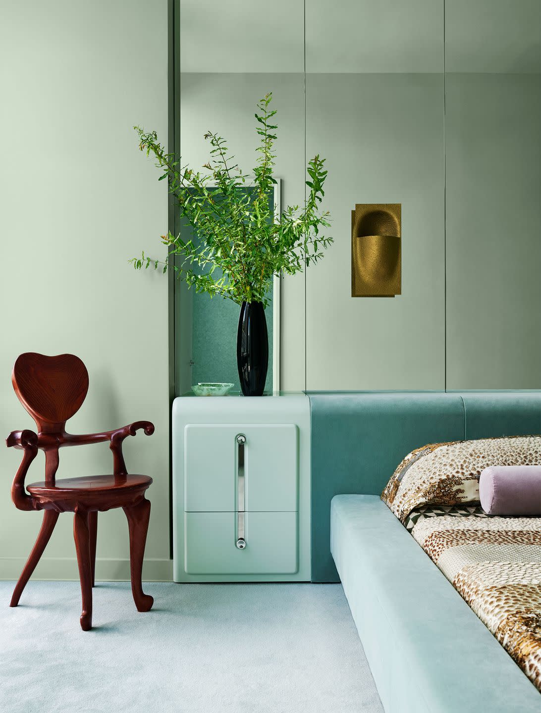 a bedroom has mint green walls, a mirrored wall behind a bed upholstered in a light aqua velvet and a light green nightstand, bronze sconce, an oak armchair with a heart shaped back, mint green carpet