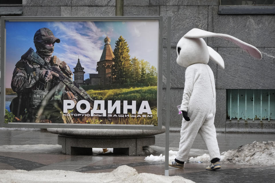 A street performer calling people to visit a museum walks past an image of a Russian serviceman reading 'The Motherland we defend' at a street exhibition of military photos in St. Petersburg, Russia, Tuesday, March 14, 2023. (AP Photo/Dmitri Lovetsky)