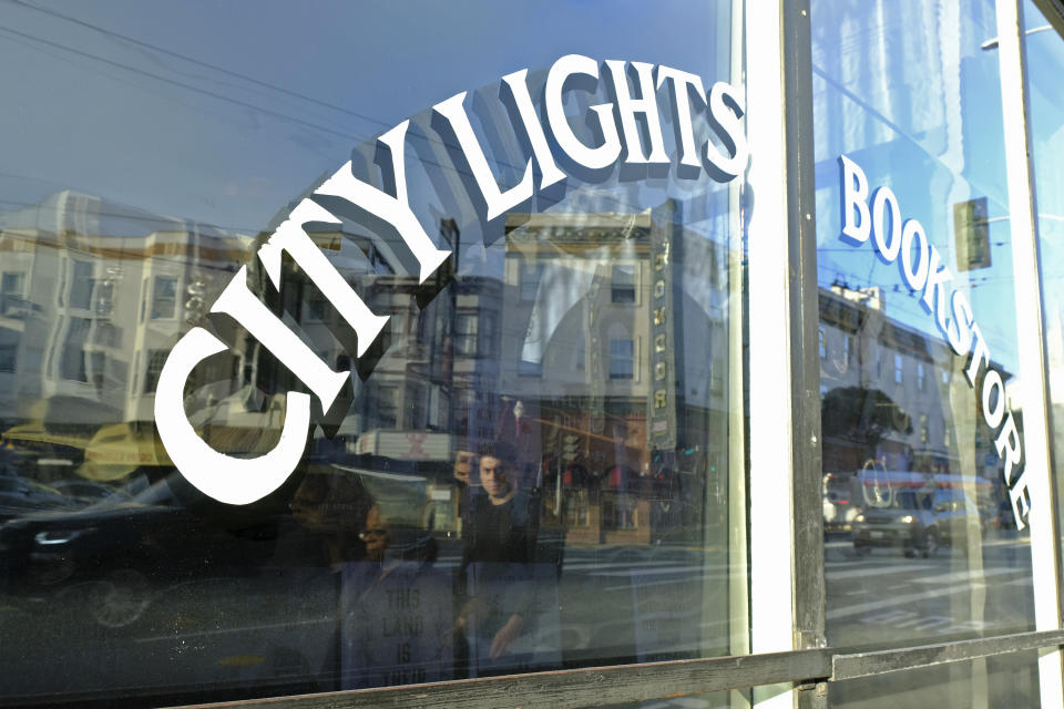 FILE - In this March 13, 2020 file photo, people are reflected in the window of the City Lights Bookstore in North Beach as they walk along Columbus Avenue in San Francisco. The famous independent bookseller was in dire financial shape because of the coronavirus outbreak and asked for help. Within days of starting a GoFundMe campaign last week seeking $300,000, the store received more than $400,000, from thousands of contributors. (AP Photo/Eric Risberg, File)