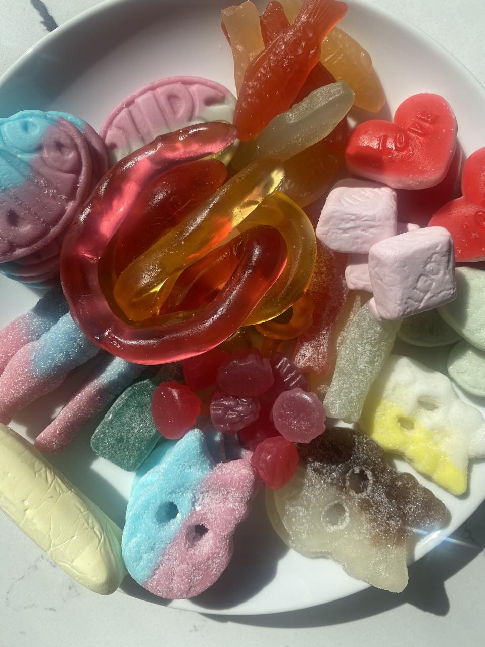 Assorted gummy candies in various shapes and flavors on a white plate. The candies include gummy worms, hearts, skulls, and more. The plate is on a light background