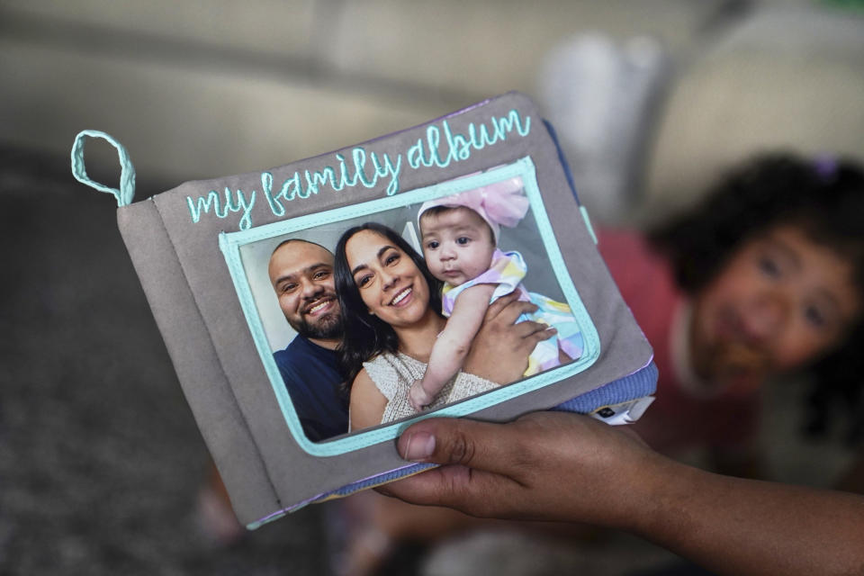 Laura Guerra gives her daughter Emilia, 2, a soft book with her father's pictures at their home in Riverside, Calif., on Monday, July 11, 2022. For now, Laura is focused on making sure her daughter, Emilia, remembers her father. Rigo Guerra passed away on Dec. 24, 2020, due to complications following a monthlong battle with COVID-19. California has approved trust funds for some children from low-income families who lost a parent or caregiver to COVID-19. The Legislature set aside $100 million in the state budget to put into trust funds. (AP Photo/Damian Dovarganes)