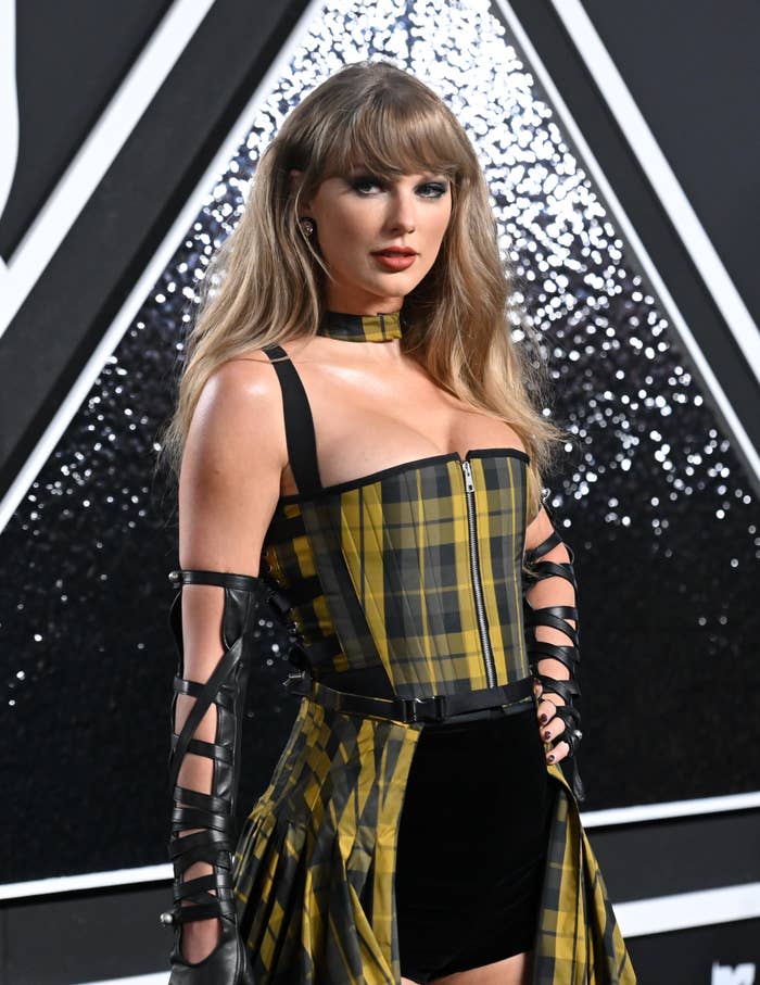 Taylor Swift wears a black and yellow checked corset dress with a flared skirt and long black ribbons and poses in front of a patterned background