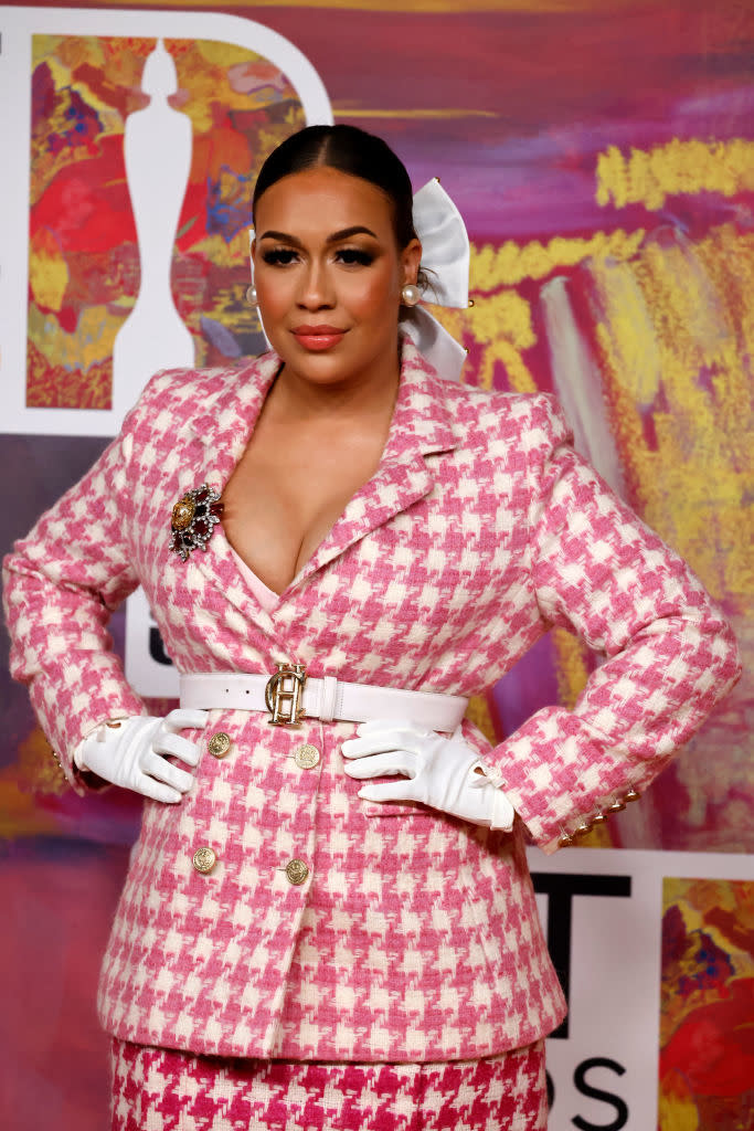 A woman poses confidently at an event, wearing a stylish pink and white patterned suit, matching skirt, white gloves, and a large white bow in her hair