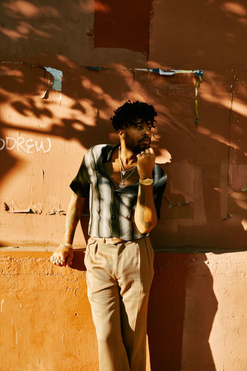 A man stands outdoors in a mix of sun and shadow against a wall.