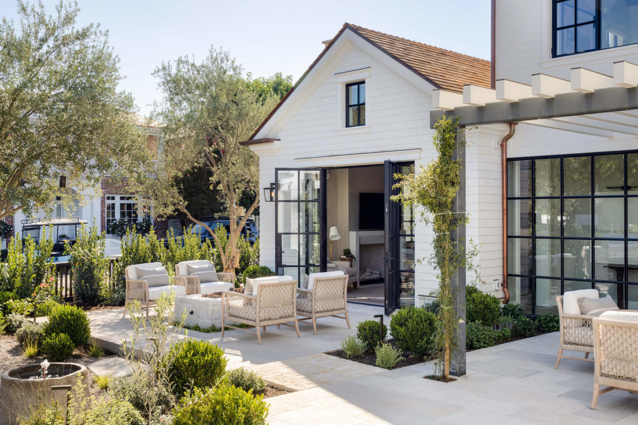  A sunny patio area with furniture, evergreen plants and a white house. 