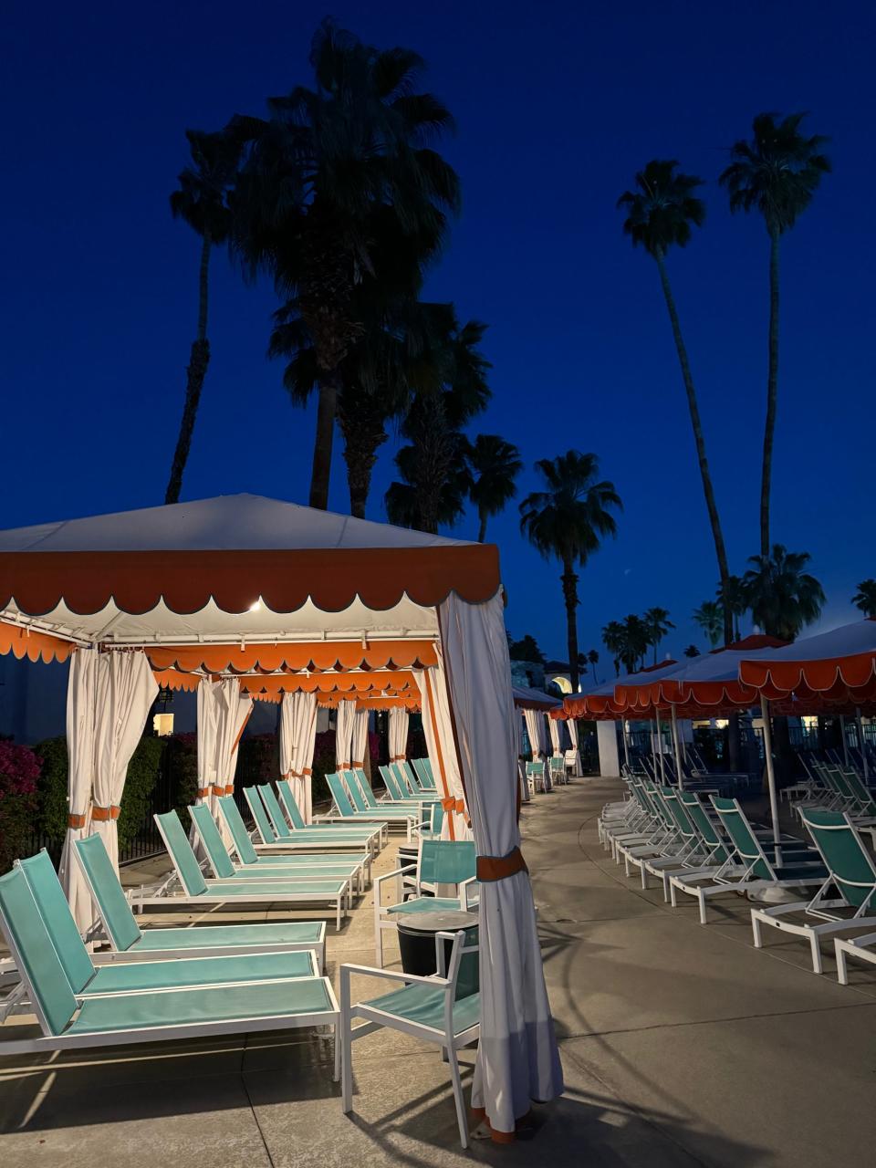 Poolside loungers and cabanas at a resort during twilight with palm trees in the background