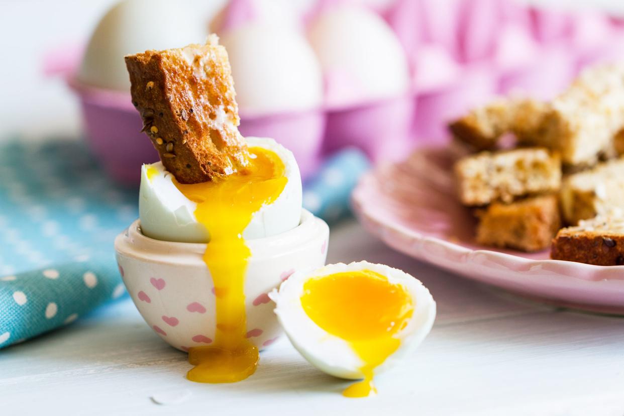 A soft-boiled egg and soldier in a pink heart egg dish, next to a pink plate of soldiers, and eggs in the background