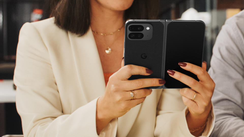 Product lifestyle image for the Pixel 9 Pro Fold. A person holds a foldable phone (open) next to another person in a modern, open conference area in a business environment.