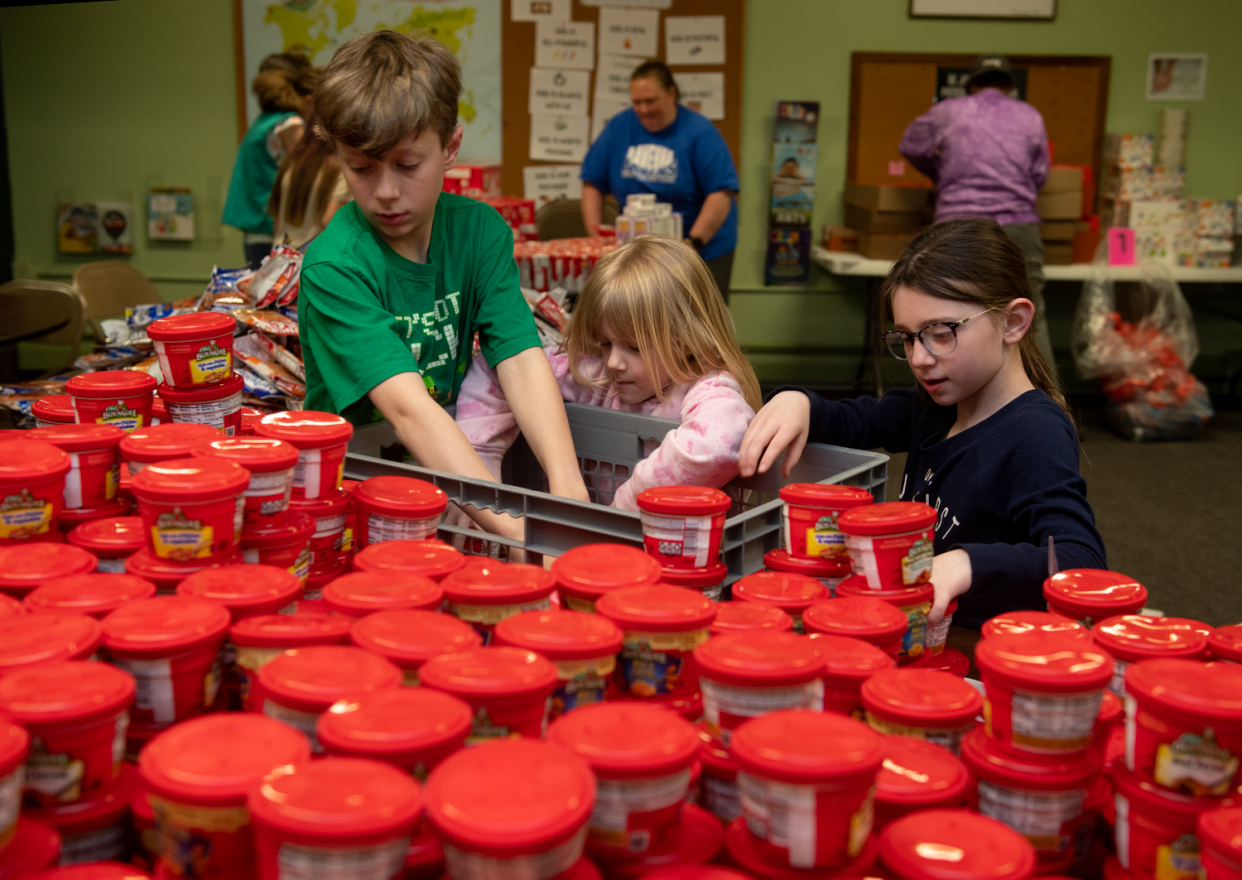 The Wisniewski family — Mikey, 10, Lily, 5, and Genevieve, 7 — unpack microwave dinners while volunteering for Raven Packs on April 27.