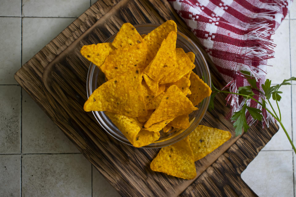 A glass bowl filled with tortilla chips is placed on a wooden board, beside a red checkered cloth and a sprig of parsley