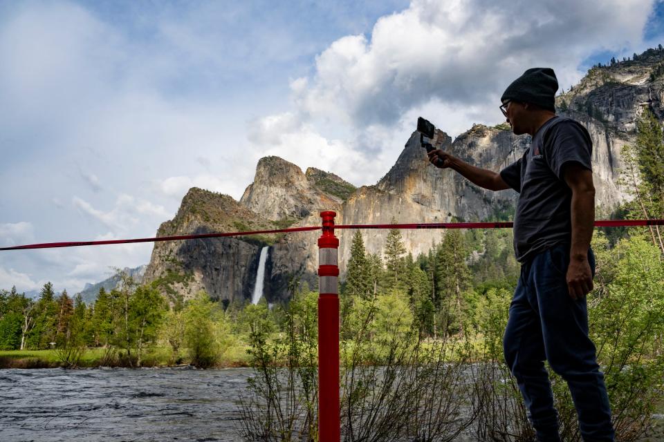 A man records Thursday, May 25, 2023 behind a temporary barrier in Yosemite National Park as Bridalveil Fall runs in the distance. Significant flooding has altered many areas.