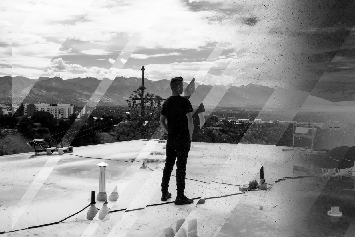 Black and white image of a man in a black T-shirt, standing on a roof and facing away from the camera toward a mountain vista.
