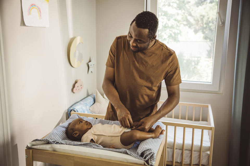 Man changing a baby's clothes in a crib, indoors, next to a window