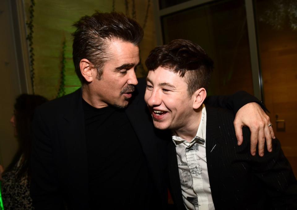 colin farrell and barry keoghan stand next to own another in a dark room, farrell looks at and talks to keoghan with one arm around his shoulder, keoghan smiles