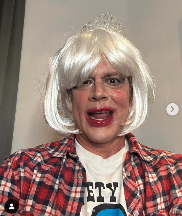 Person in platinum wig and tiara, wearing plaid shirt over graphic tee, with red lipstick