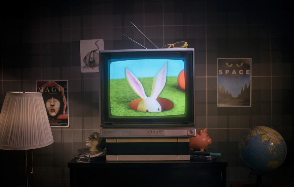 A retro-styled living room with a TV showing a cartoon rabbit peeking from a hole. Surrounding the TV are posters, a lamp, a globe, and a piggy bank