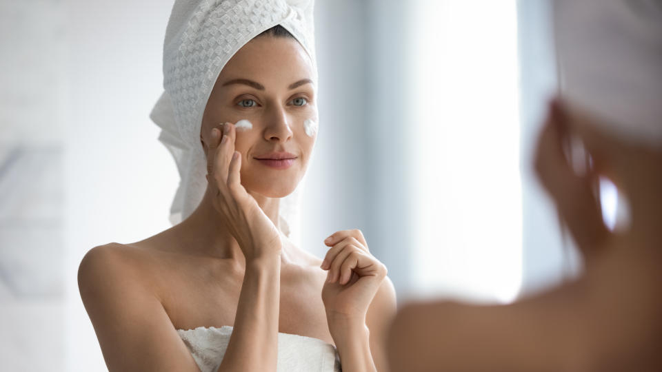 Attractive young adult woman apply facial cream look in mirror, beautiful healthy lady wrapped in towels put moisturizing lifting nourishing day creme on soft hydrated moisturized skin in bathroom