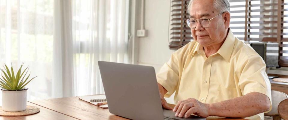 Older man types on laptop, sitting at kitchen table