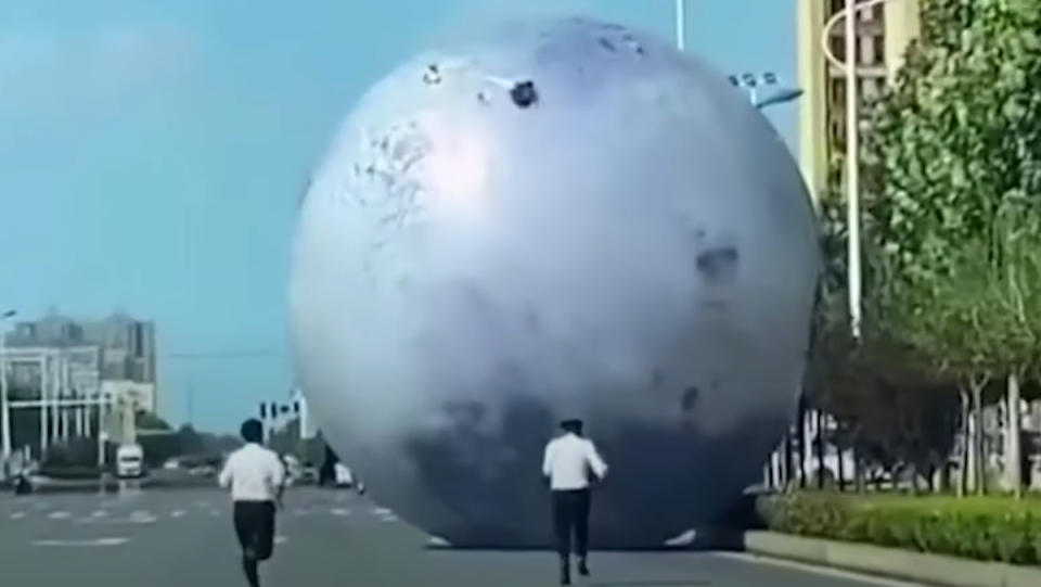 A giant, inflatable Moon rolling down the road as two men chase after it on foot.