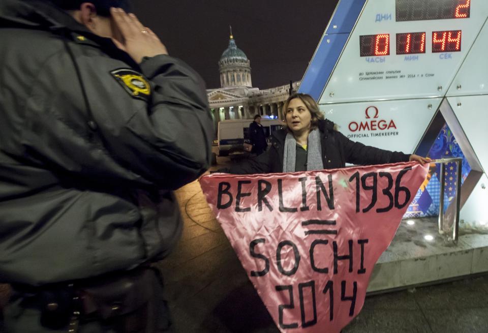 A gay rights activist holds a banner in front of a large clock showing the number of days left until the start of the Olympic games as a police officer approaches, left, in St. Petersburg, Russia, Wednesday, Feb. 5, 2014. Russian gay rights activists protested the upcoming Olympic Games in Sochi. Two activists unfurled banners reading “Berlin 1936 = Sochi 2014,” referring to the Olympic Games that were held in the capital of Nazi Germany. One-man pickets are legal in Russia and the two activists holding signs were spaced far enough apart that neither was arrested. (AP Photo/Elena Ignatyeva)