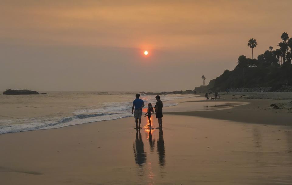 Beach-goers wade in the water as the sun and sky are partially obscured with ash and smoke.