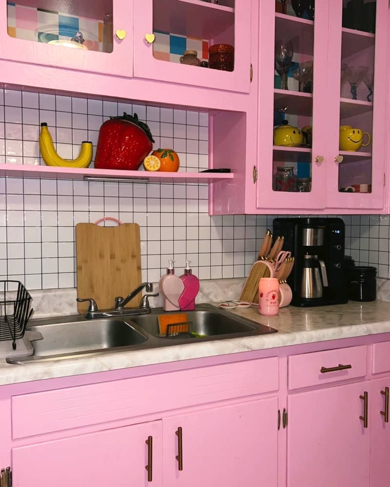 Fruit decor on shelf above double sink in kitchen with white tile backsplash and pink cabinets.