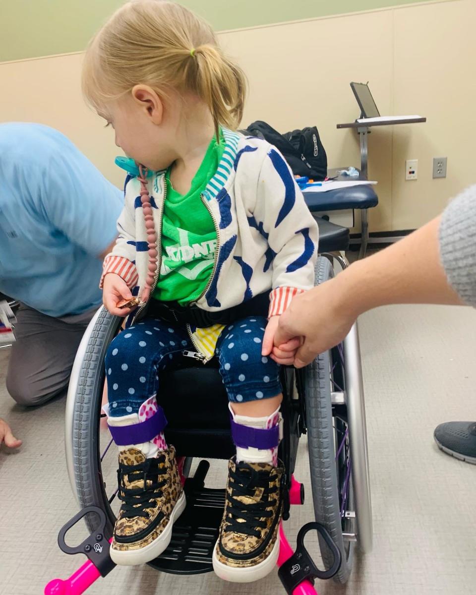 Katie Spence's daughter in a wheelchair wearing leopard print shoes and a colorful cardigan.