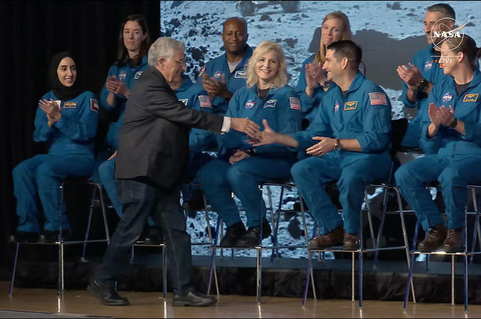 a man in a dark suit shakes hands with a seated man in a blue flight suit while other people in blue flight suits smile