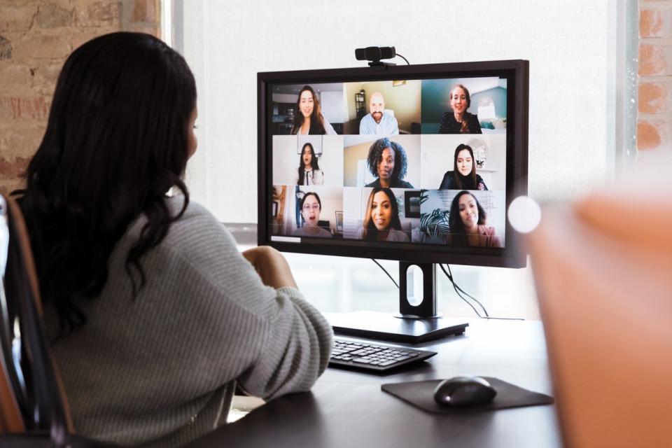 A professionally dressed work-from-home employee speaking to her coworkers on a video call using a desktop computer and webcam.