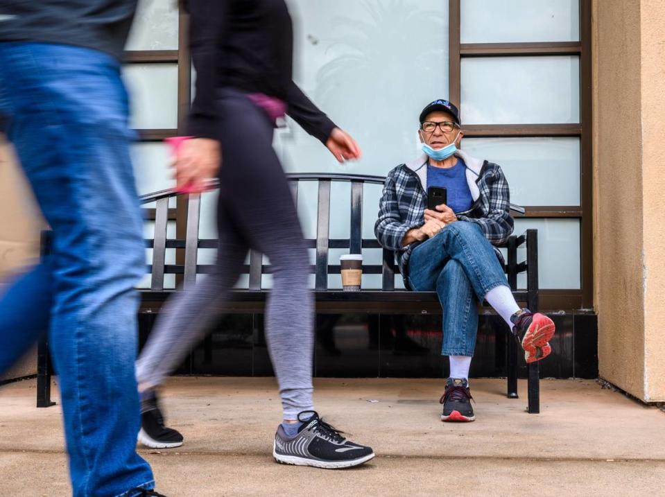 Carmelo Di Grande sits on a bench, mask at ready, with a beverage as people walk past him at Fountains at Roseville on Monday, May 18, 2020.