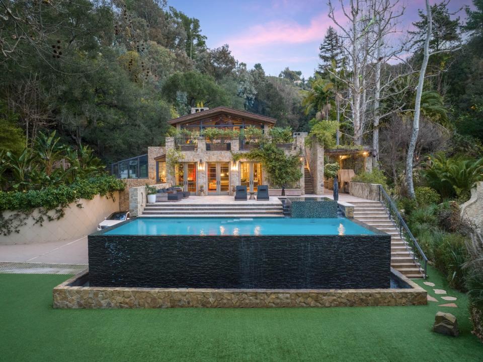 An elevated infinity pool sits in front of what is the back of the multi-story home that is surrounded by trees