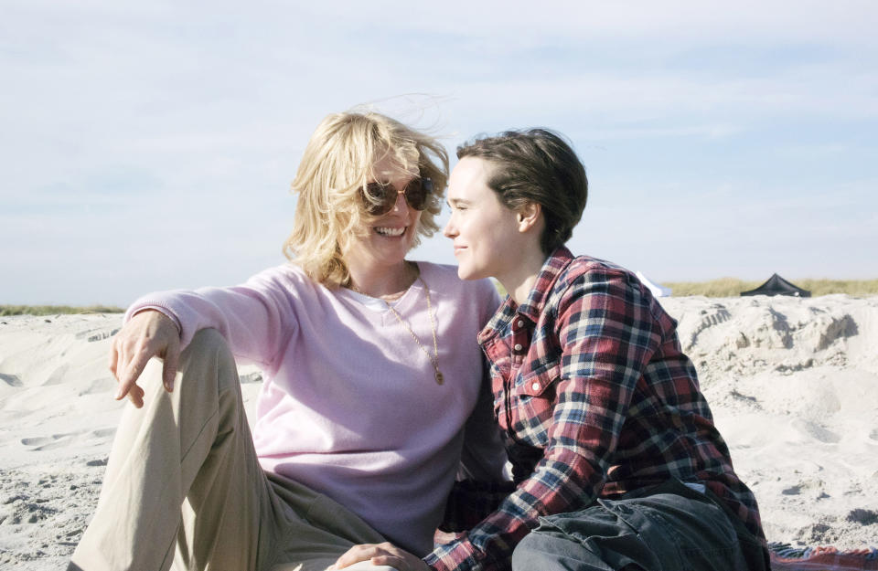 Julianne Moore and Elliot Page sit on a beach, smiling at each other, with Julianne wearing a light sweater and sunglasses, and Elliot in a plaid shirt