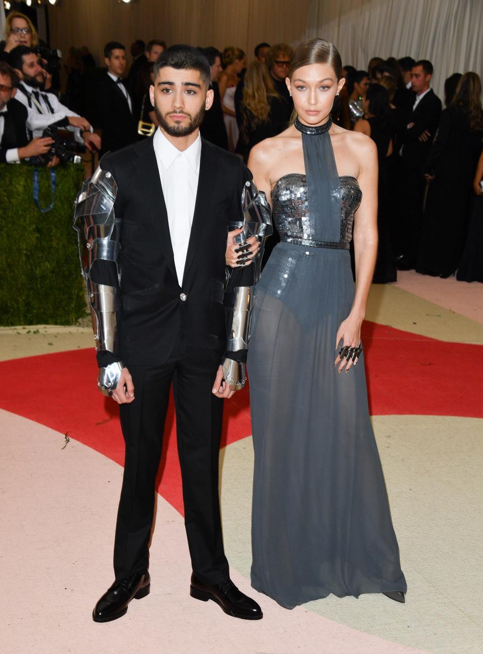 Zayn Malik stands in a suit and Gigi Hadid holds his arm wearing a grey dress on a red carpet.