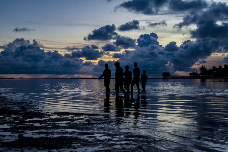 A group of people cross a shallow lagoon at dusk in the tropics.